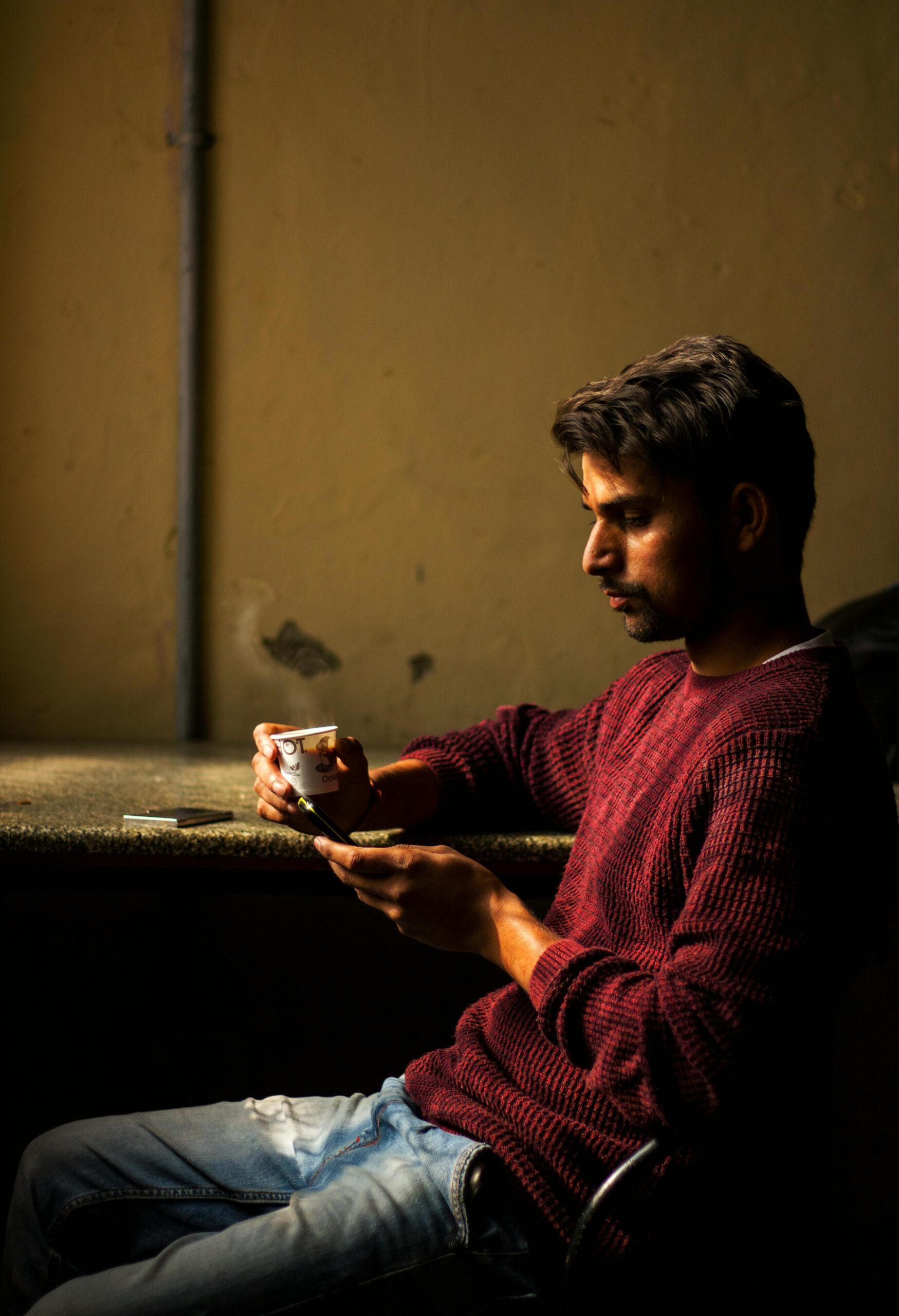 A young man in a maroon sweater sits indoors, holding a cup and using a phone.
