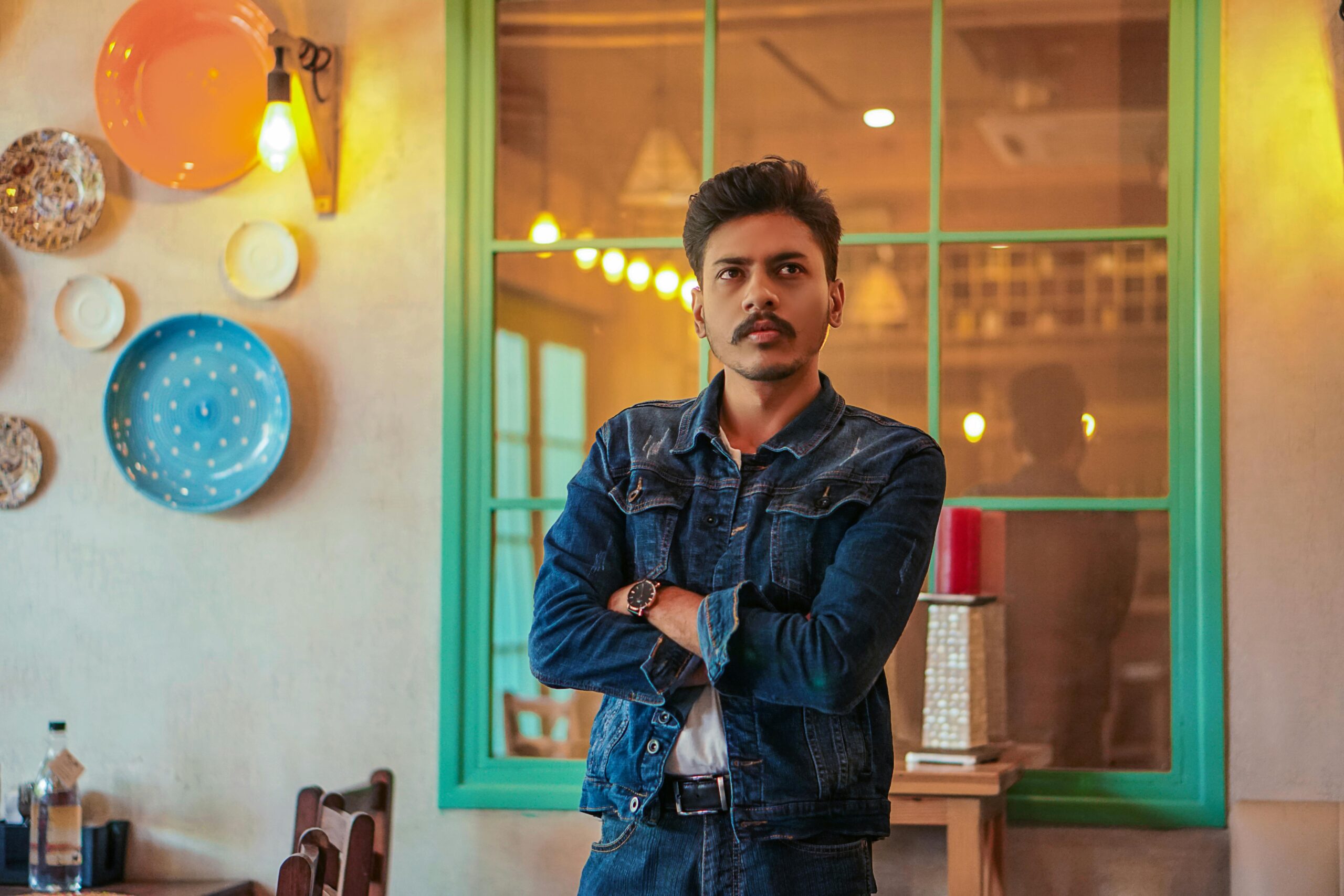 Young man with mustache in a denim jacket posing indoors with reflective window and wall decor.