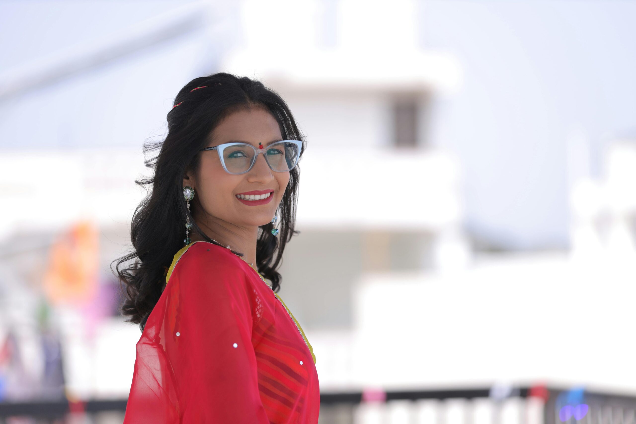 Smiling woman in a vibrant red saree posing outdoors, showcasing traditional Indian fashion with modern eyewear.