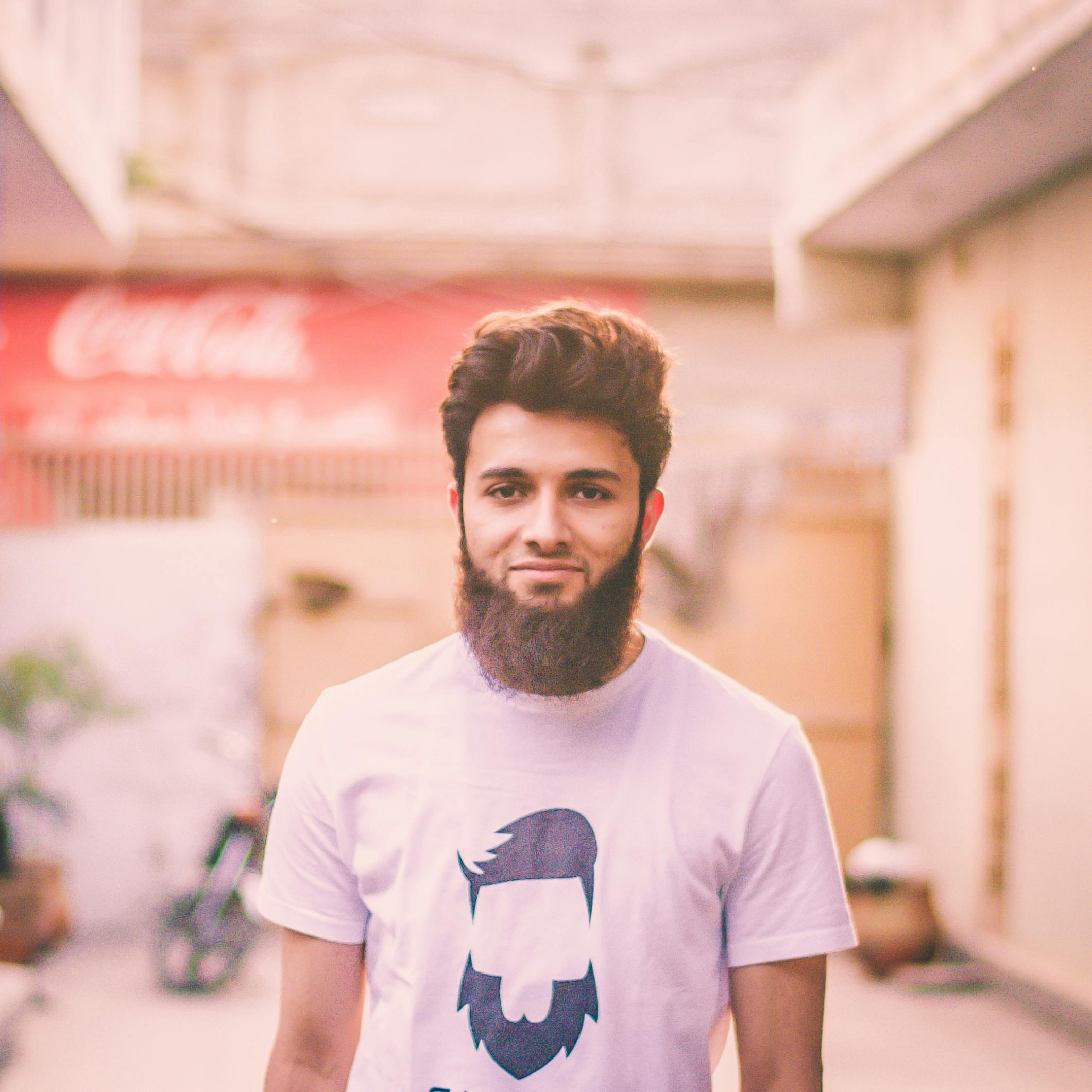 A young bearded man smiling warmly, dressed casually in a t-shirt, captured outdoors in a soft light.